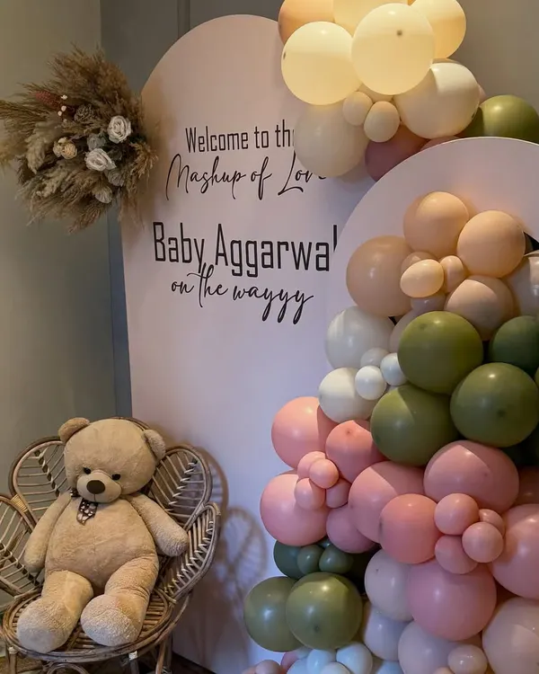 "Welcome to the Mashup of Love." For this baby shower, we designed a backdrop with soft, earthy tones of pink, olive green, and cream balloons, accented with a dried floral bouquet and a cute teddy bear on a wicker chair.