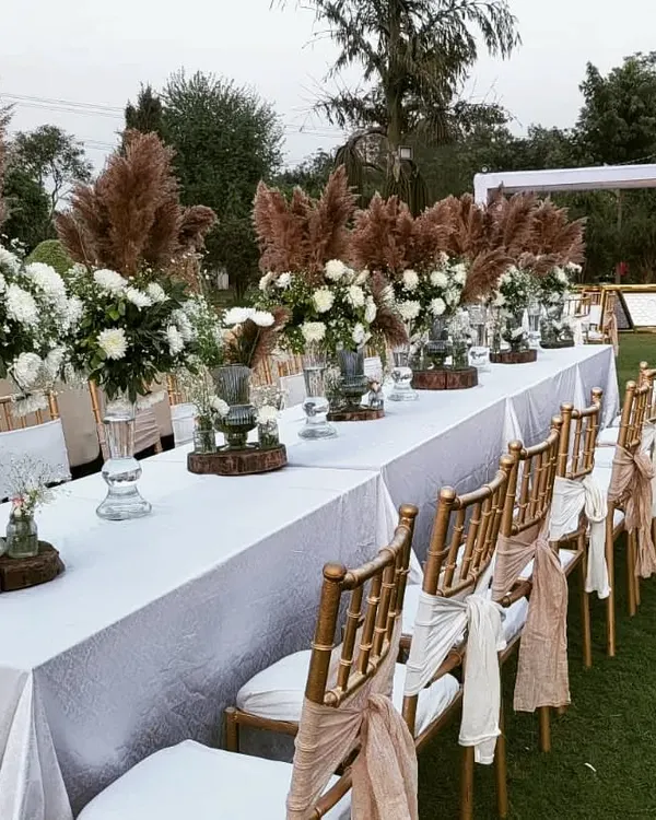 An elegant table runner for a Boho-themed wedding. The long table is decorated with pampas grass, white roses, and wooden log slices for a rustic yet chic look.