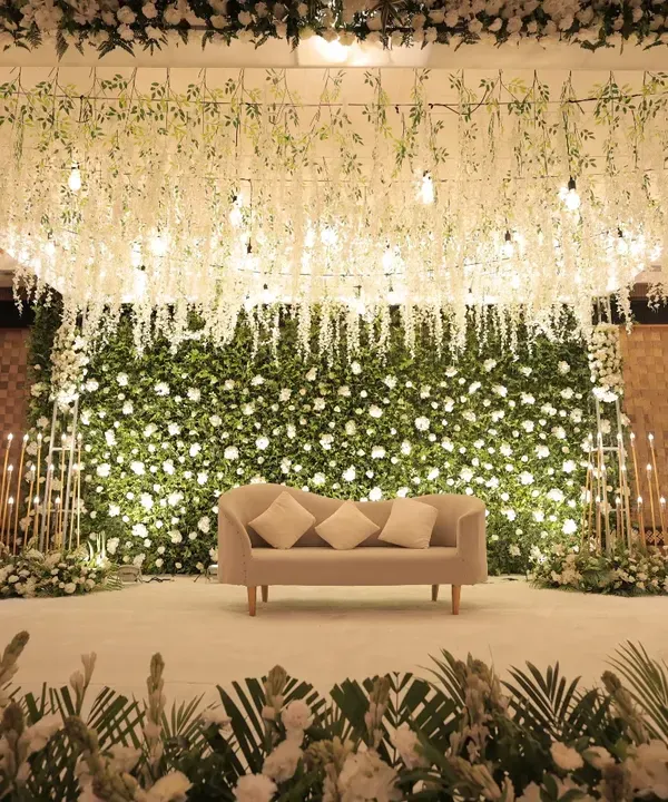 A direct view of our magnificent white floral stage. The design features a dense wall of white roses as a backdrop, with a ceiling of hanging wisteria and delicate lights, creating a breathtaking setting for a wedding or reception.