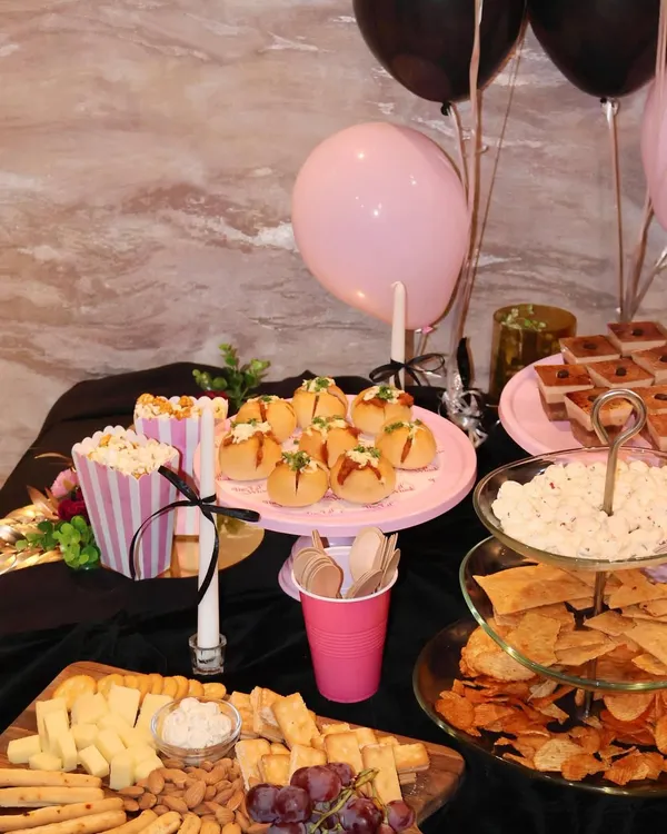 A complete party table setup featuring a mix of sweet and savory options. You can see our pull-apart breads on a pink stand, finger sandwiches, a cheese board, and various dips, all coordinated for a chic black and pink theme.