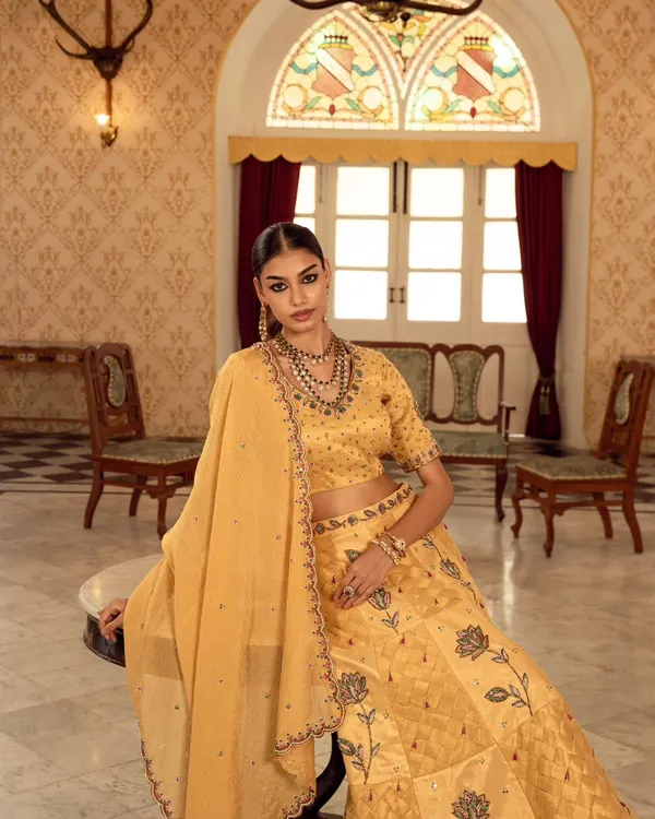 A moment of quiet elegance captured for a luxury ethnic wear campaign. The soft, natural light from the arched window illuminates the detailed embroidery on the yellow lehenga.
