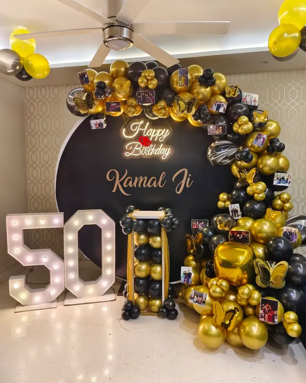 A personalized 50th birthday party featuring a black and gold balloon arch decorated with photos, a neon sign, and large '50' marquee numbers.