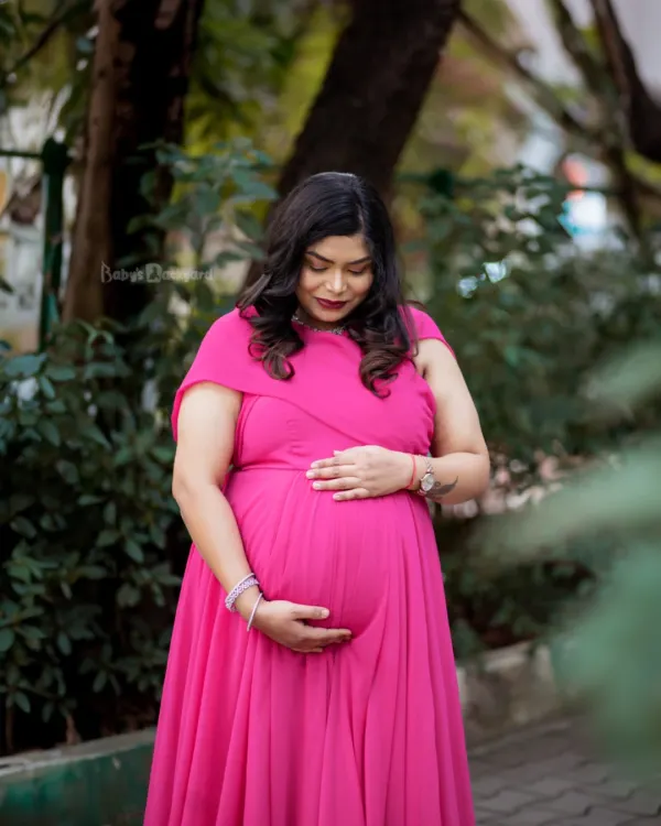 A moment of quiet reflection for the mom-to-be. Dressed in a vibrant pink gown, she gently holds her bump, surrounded by the soft light of an outdoor setting.