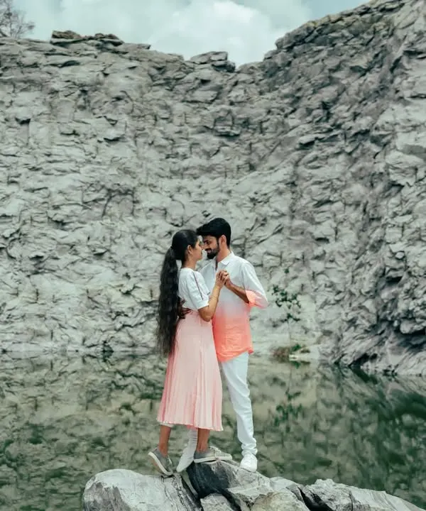 A stunning wide shot of a couple standing on a rock by a tranquil lake, framed by massive rock formations. The scale of the landscape makes this a truly epic pre-wedding photo.