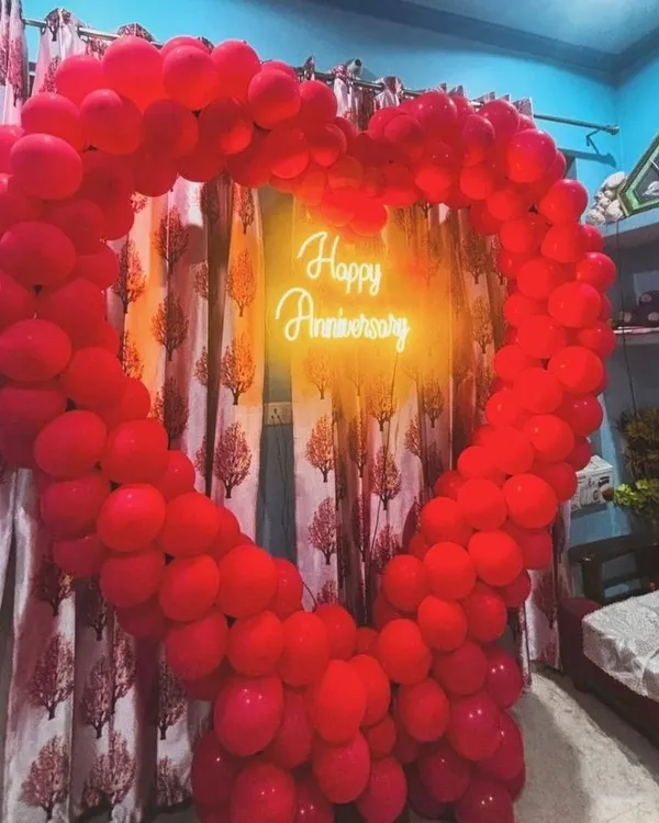 A romantic heart-shaped balloon decoration for a wedding anniversary at home. The frame is made of red balloons with a 'Happy Anniversary' neon sign inside.