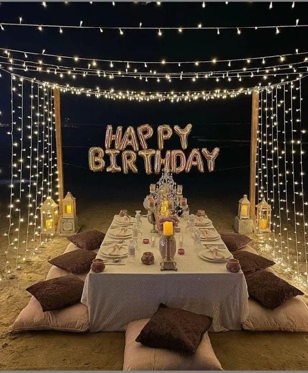 A dream-like birthday dinner setup on the beach. We created a canopy of fairy lights over a low-seating dining table, with "Happy Birthday" foil balloons and lanterns creating a magical atmosphere.