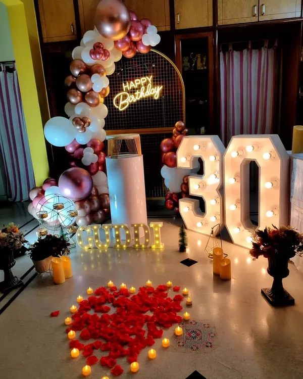 A chic 30th birthday celebration in a modern home. The decor features large light-up marquee numbers, a rose gold balloon arch, and a heart made of petals and candles on the floor, creating a stylish and personal atmosphere.