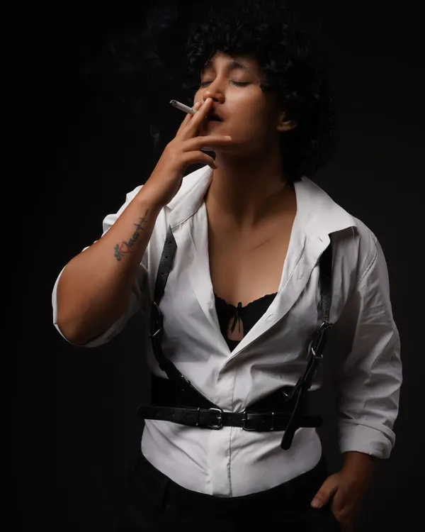A moody studio portrait from the "Leather straps and Cigarettes" series. The dark background and single light source create a dramatic, film-noir atmosphere.