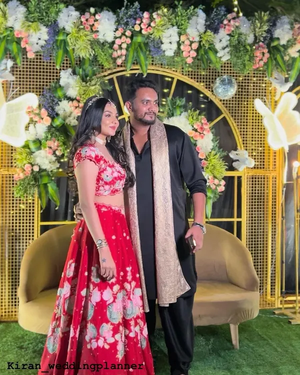 A stylish couple posing at their Sangeet night. The backdrop features a modern gold grid decorated with flowers and glowing butterfly motifs, creating a chic and festive vibe.