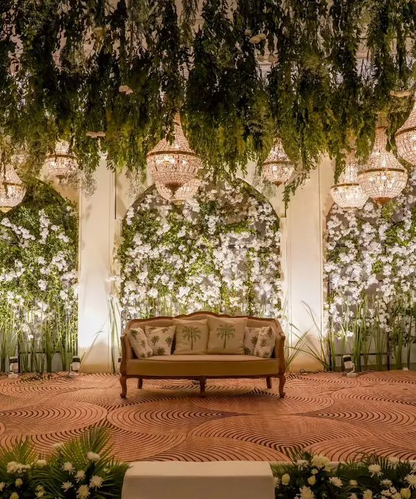 This reception stage combines a lush overhead canopy of greenery and crystal chandeliers with dense floral walls, creating a breathtaking focal point for the evening's celebrations.