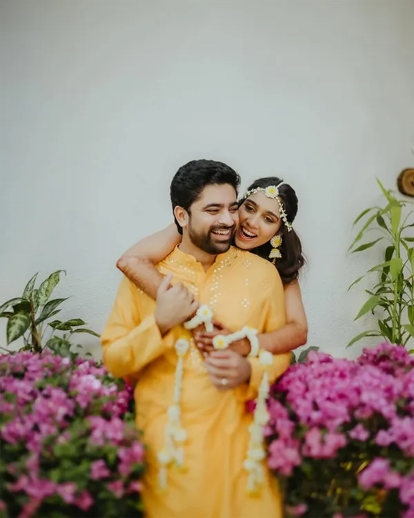 A joyful and candid moment from a Haldi ceremony, with the bride-to-be playfully hugging her partner from behind amidst a backdrop of beautiful purple flowers.
