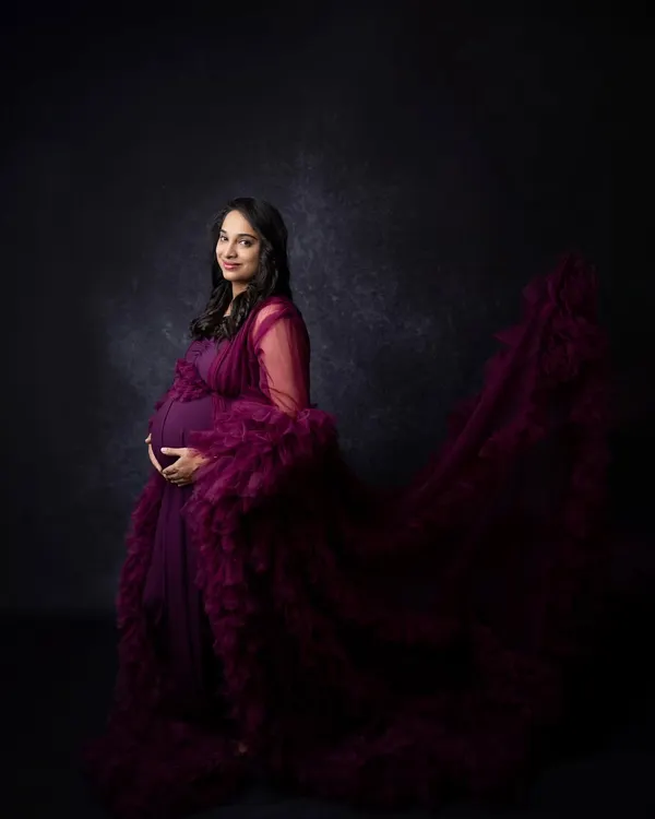 This dramatic maternity portrait uses a deep purple gown with a long, flowing train against a dark, textured backdrop to create a powerful and artistic image. The lighting is carefully shaped to highlight the mother's form and the richness of the fabric.