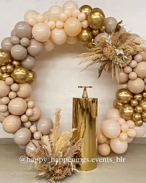 A full circular balloon arch makes a spectacular photo backdrop. This design uses a sophisticated palette of beige, brown, and chrome gold, accented with dried florals and pampas grass for a modern, earthy feel.