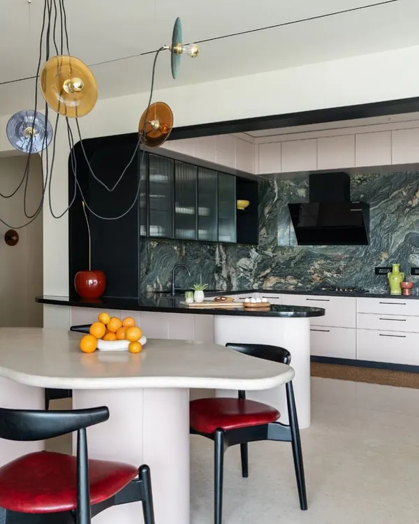 This open-plan kitchen and dining area was designed to be bold, eccentric, and fun. An amorphous, custom-cast dining table flows into the kitchen island, while a dramatic green marble backsplash and colorful pendant lights add pops of color and playfulness.