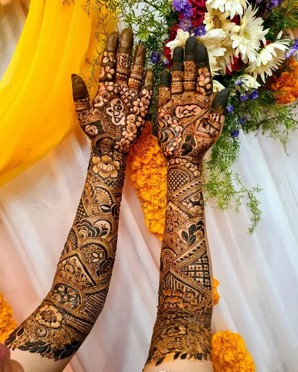 Full-arm bridal henna with a stunning contrast of bold and fine patterns. The design features geometric shapes, paisleys, and floral elements, set against a festive backdrop.