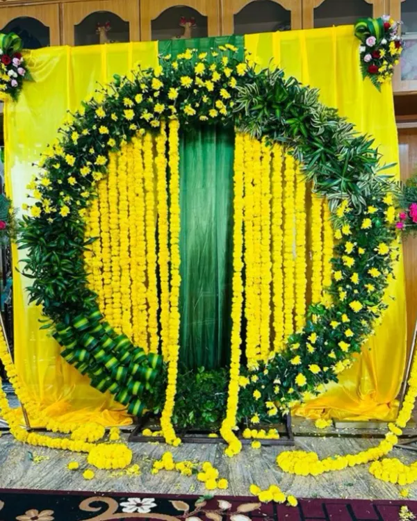 A classic Haldi ceremony backdrop, full of vibrant yellow. This design features a large circular arch made of lush green leaves and yellow flowers, with strings of marigolds hanging in the center against a yellow and green drape.
