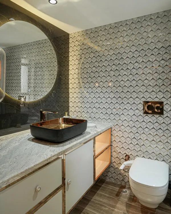 A modern washroom with geometric 3D wall tiles, a black and gold vessel sink, and a large round backlit mirror.