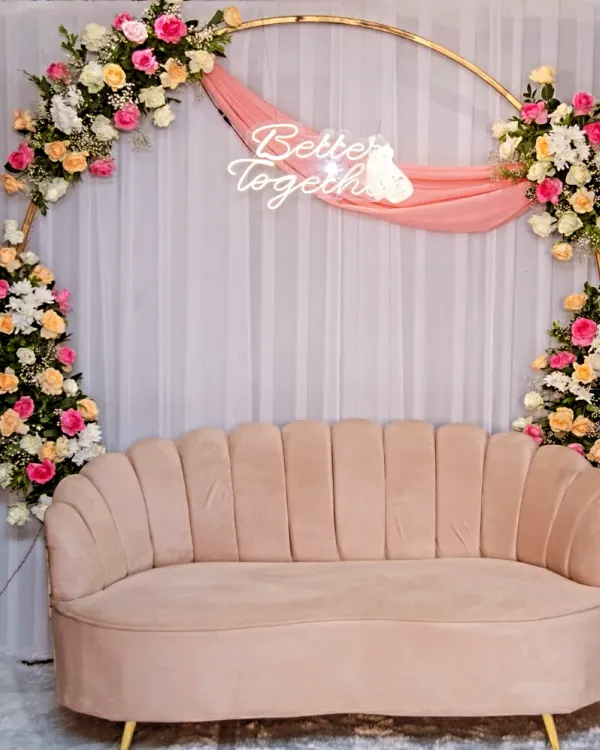 A lovely seating area for the happy couple. This setup features a modern pink velvet sofa in front of a circular golden arch decorated with pink, peach, and white flowers and a "Better Together" neon sign.