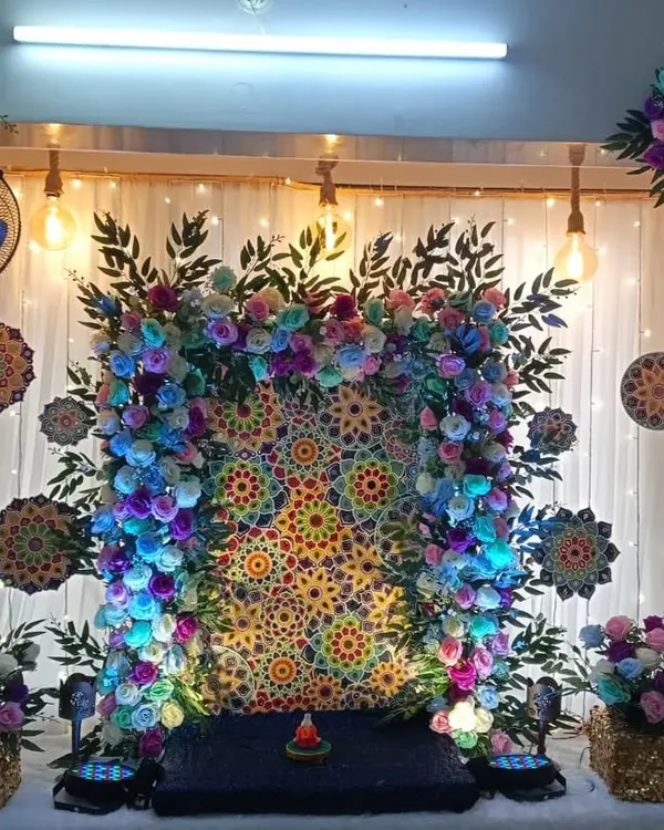 A vibrant home Ganpati decoration with a colorful mandala-style backdrop. The setup is framed by a floral arch and illuminated with blue light for a divine effect.
