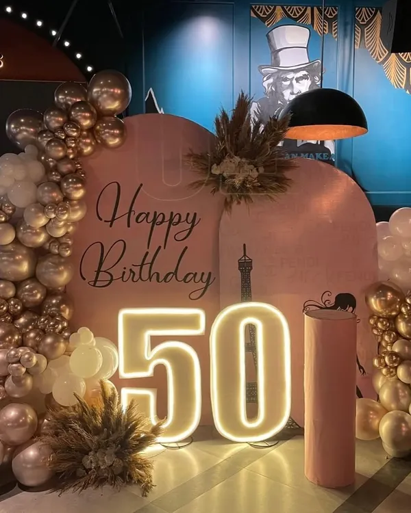 A chic and modern setup for a fabulous 50th birthday. This design features a double U-shaped blush backdrop, a stylish organic balloon arch in champagne gold and white, and dried pampas grass for a touch of boho elegance, all illuminated by a large "50" LED number.