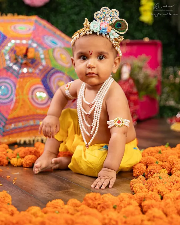 Our little Krishna, looking so regal with his pearl necklaces and crown. The colorful backdrop and marigold flowers create a vibrant, festive atmosphere.