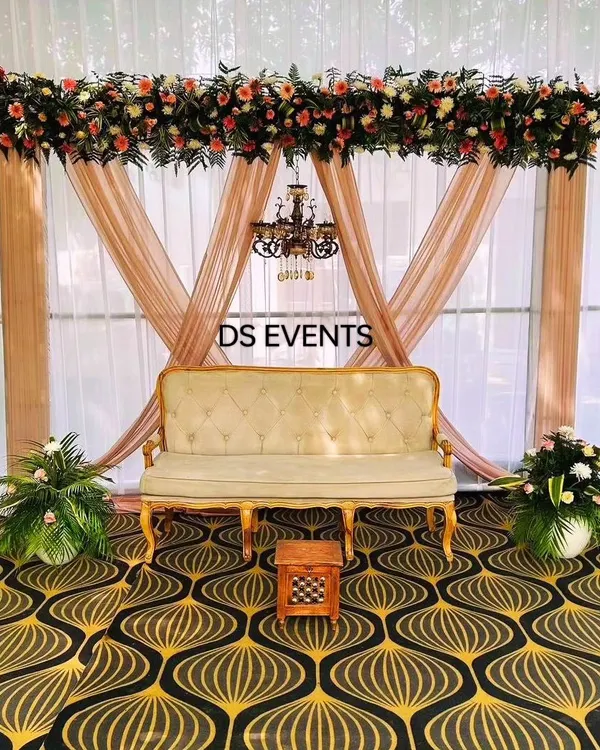 A classic and elegant stage setup. This design features soft drapes, a beautiful floral arrangement overhead, a vintage-style chandelier, and a royal sofa, perfect for a reception or anniversary celebration.