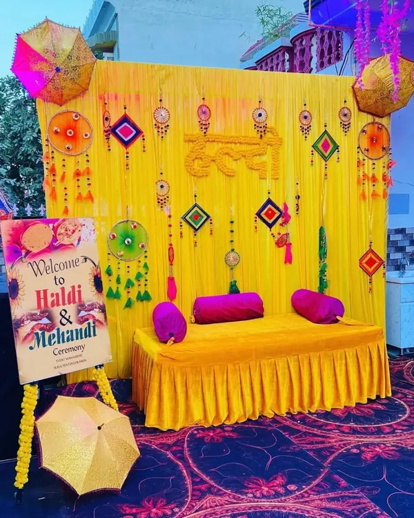 A complete Haldi and Mehendi ceremony setup with a decorated seating bench. The backdrop is a vibrant yellow fabric adorned with traditional hangings, flanked by Rajasthani umbrellas and a custom welcome sign for the event.