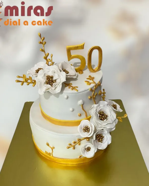 A stunning two-tier cake for a golden 50th birthday. The elegant white and gold design is adorned with beautiful, handcrafted white and gold sugar flowers, creating a sophisticated and memorable centerpiece.
