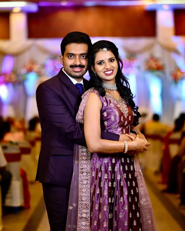 A classic posed portrait of a couple during their wedding reception. The groom is in a sharp suit and the bride wears an elegant purple lehenga, perfectly captured against the stage decor.