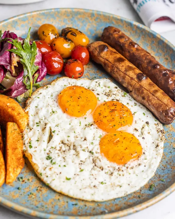A classic breakfast plate featuring sunny-side-up eggs with perfectly runny yolks, paired with savory sausages, potato wedges, and a side of fresh greens. This is the kind of meal that fuels your entire day.