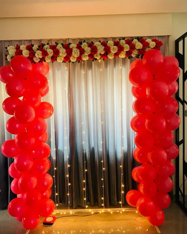 This romantic backdrop features two bold pillars of red balloons, topped with a garland of red and white artificial flowers. The fairy lights on the curtain behind add a soft, warm glow to the setting.