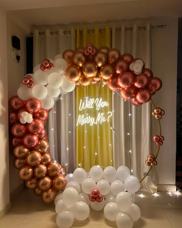 A beautiful indoor proposal setup. This features a large circular ring decorated with a cascade of metallic rose gold and white balloons, with a 'Will You Marry Me?' neon sign in the center.