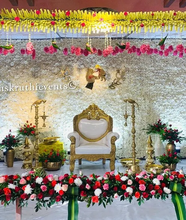 An elegant and unique stage for a traditional ceremony. The backdrop is a dense wall of white jasmine, framed by columns of flowers and a border of red and white roses.