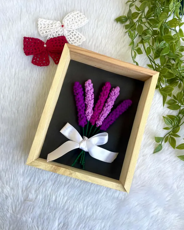 A beautiful arrangement of crochet lavender sprigs, tied with a white ribbon and set inside a wooden frame. This is a simple yet elegant piece of floral wall decor.
