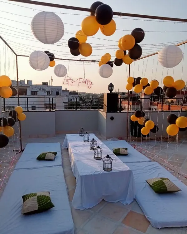 A relaxed and stylish terrace party setup. This decoration features a canopy of fairy lights and paper lanterns, with low seating arrangements and a 'Let's Party' neon sign.