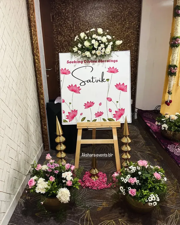 A beautiful welcome sign for a naming ceremony, featuring a lotus theme. The sign is flanked by traditional brass lamps and pots of fresh flowers, creating an auspicious entrance.