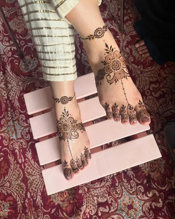 A beautiful and delicate foot mehendi design, featuring a central floral motif with a trailing pattern on the toes and a matching anklet band.