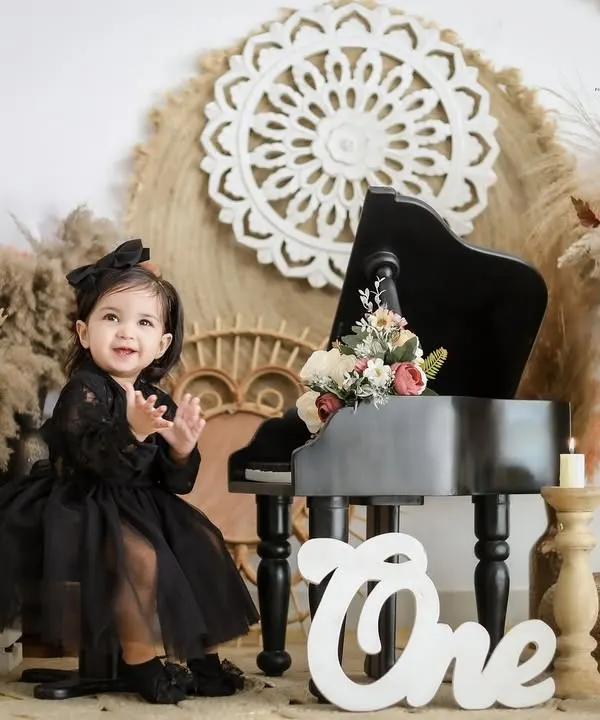 A little star claps her hands with excitement during her boho themed first birthday shoot. The setup includes a miniature piano, pampas grass, and a 'One' sign for a chic and stylish look.