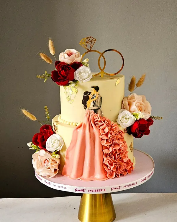 An absolutely stunning engagement cake. This design features a beautiful couple topper with a flowing, ruffled dress, surrounded by handcrafted sugar flowers in shades of red, pink, and white.