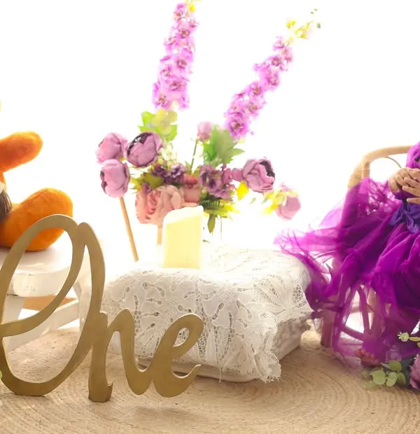 Celebrating her first birthday with a smile and a tea party. This little girl in a beautiful purple dress is surrounded by flowers and her teddy bear companion, marking her special milestone in style.