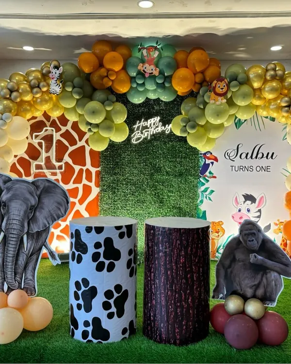 A detailed jungle safari birthday stage. This decoration features a grass wall, animal print pedestals, large cutouts of an elephant and gorilla, and a multi-toned balloon arch for a complete jungle experience.