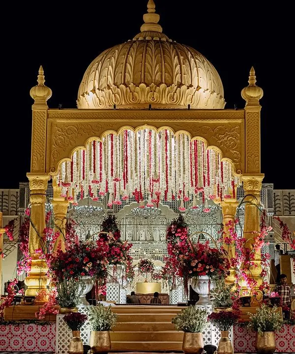 The main wedding mandap, a magnificent golden structure with a central dome and cascading floral garlands. It stands as a beacon of royal elegance against the night sky.