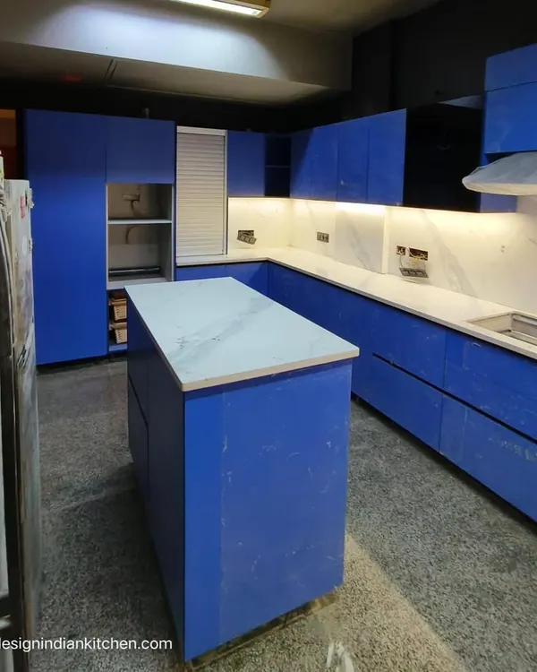 A stunning L-shaped kitchen featuring bold, matte blue Fenix shutters, currently under installation in Vasant Vihar. The design includes a central island and is paired with a white marble-patterned countertop for a striking contrast.