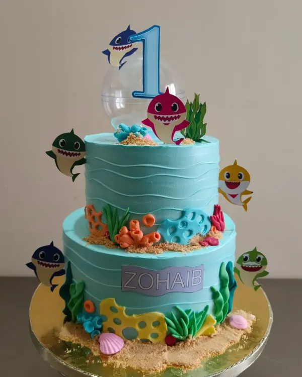 A vibrant two-tier Baby Shark cake, perfect for a first birthday. This underwater scene is created with blue buttercream waves, edible sand, and all the favorite shark characters.