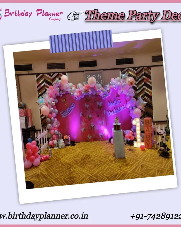 A full view of a pink-themed first birthday party. The decor includes a custom backdrop with balloon garlands, a 'BABY' marquee sign, and a white picket fence prop to create a defined and adorable party space.
