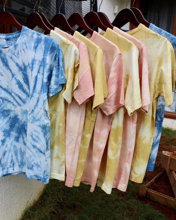 A rainbow of naturally dyed tees hanging in the sun. Each t-shirt is hand-dyed with botanical colors like indigo, madder, and marigold, making every piece unique in its pattern and shade.