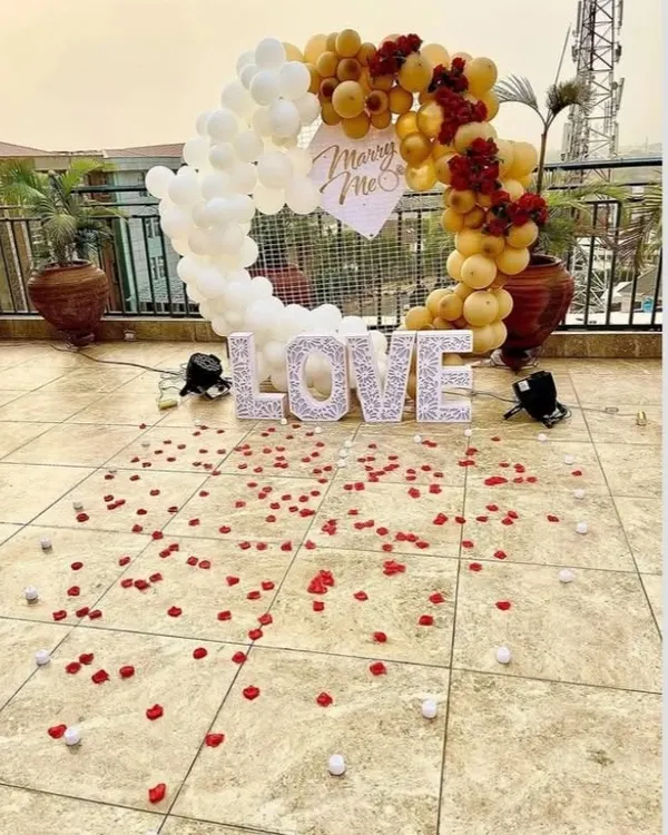 A beautiful rooftop marriage proposal setup. A white and gold balloon arch frames the moment, with 'LOVE' marquee letters and 'Marry Me' sign, all set against a city skyline.