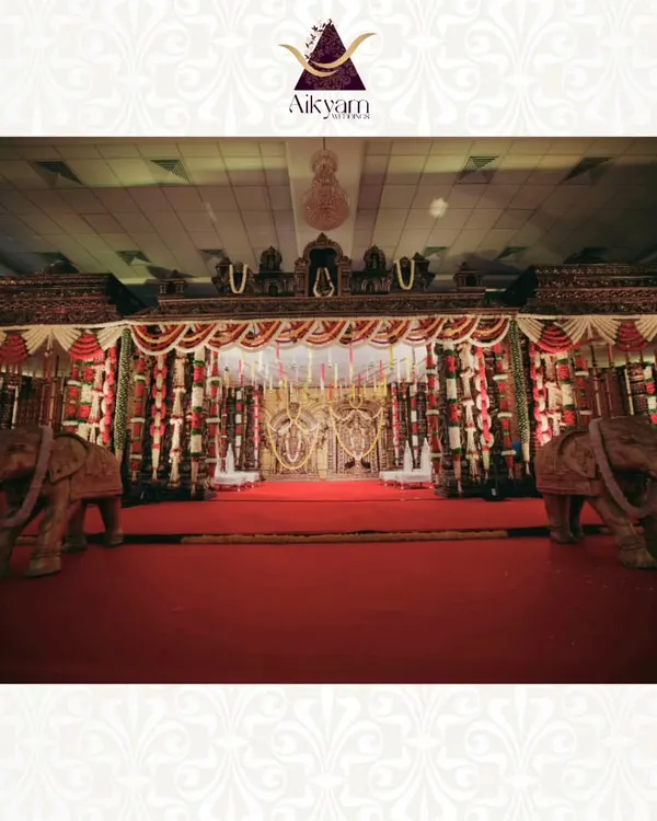 A classic temple-style mandap designed for a grand wedding ceremony. The stage is framed by carved elephants and adorned with traditional red and white floral garlands, creating a regal and sacred space.