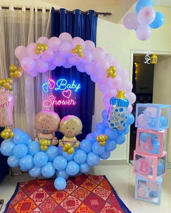 A charming baby shower decoration designed in a cradle shape using pink and blue balloons. The setup includes a 'Baby Shower' neon sign, baby-themed foil balloons, and transparent 'BABY' blocks.