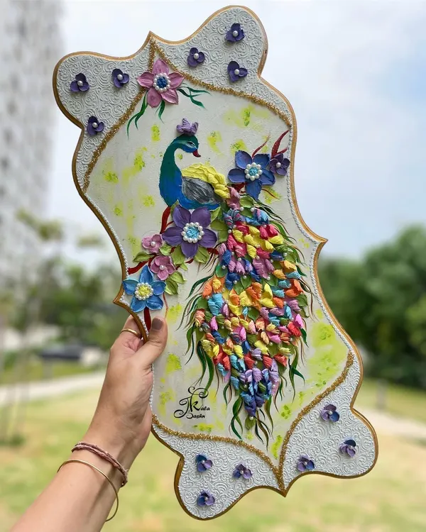 A full view of a 20-inch peacock wall art piece, held to showcase its impressive size and the cascade of multi-colored sculpted feathers. The textured white border adds an elegant finish.